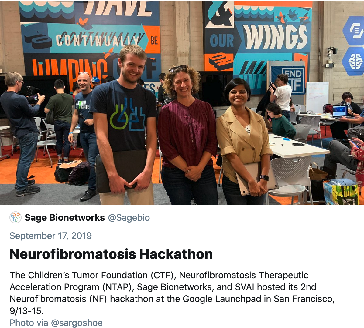 Screenshot of a group of people at a hackathon. There are colorful posters in the background.